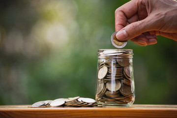 Hand of person holding a coin putting in glass jar on wooden in nature background to save money for the future plan.