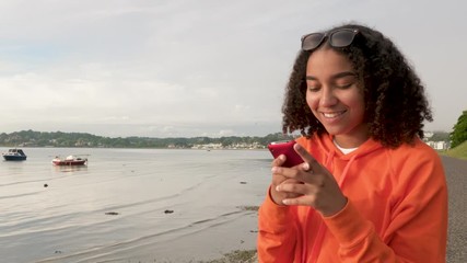 Beautiful mixed race African American girl teenager young woman wearing orange hoodie, sitting on a sea front using her smart cell phone for social media or texting