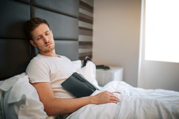 Young man fell asleep in bedroom this morning. He hold journal in hands. Daylight.