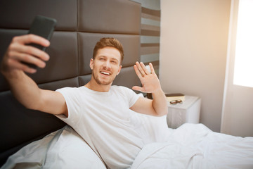 Young cheerful man in bed this morning. He and pose on phone camera. Guy wave with hand and smile. Daylight.