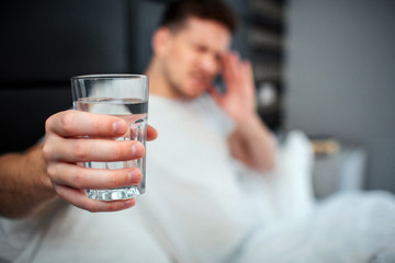 Young man in bed. He hold glass of water and show it to camera. Model hold hand on forehead. Headache or hangover.