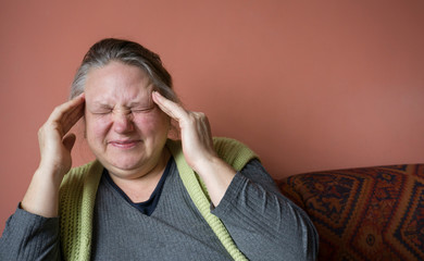 Elderly senior woman has headache. Old woman with headache. Unhappy woman in pain sitting on sofa at home, feel sick. Aged woman holding her head and suffering headache problem. Copy space for text.