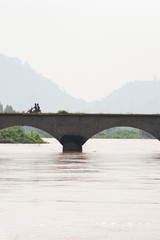 Laotians riding motorcycle crossing ancient bridge over Mekong River.