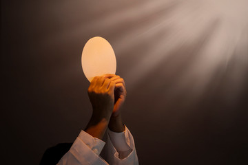 Hands of priest raise sacramental bread under light