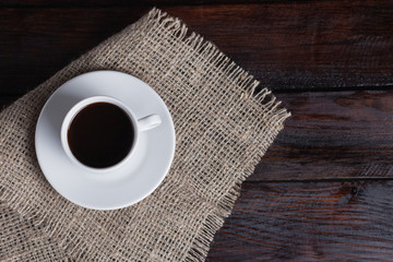 White cup of coffee on linen fabric on the dark vintage background. Top view