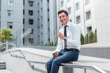 Business lifestyle. Businessman with eyeglasses sitting on fence on the city street holding cup of coffee smiling happy