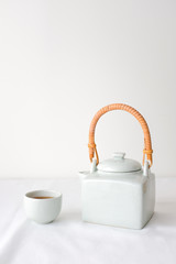 White ceramic Asian Japanese teapot with wooden handle and white ceramic teacup on white table top with white cotton fabric texture on white wall background with empty text copy space. Minimalism.