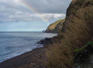 Fototapeta na wymiar Rainbow over Praia da Viola beach with grassy sandstone cliffs and volacanic rock boulders, atlantic ocean landscape, Sao Miguel island Azores island Portugal