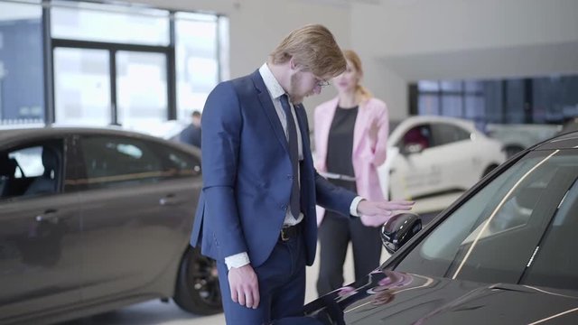 Businessman choosing the car in dealership close up. Tall man stands near automobile in auto show room, looking at car and examining vehicle, Manager shows cars to young woman in the background