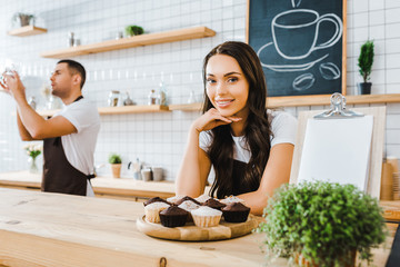 attractive brunette waitress standing behind bar with cupcakes on wooden tray wile barista holding cup in coffee house