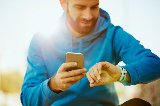 Close Up Of Young Bearded Man Looking At Smart Watch And Holding Smart Phone In Other Hand.