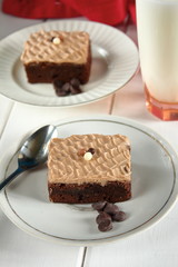 Chocolate bakery style cake brownie  with chocolate cream on top on a white plate and mint leaf and a glass of milk in the background