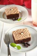Chocolate bakery style cake brownie  with chocolate cream on top on a white plate and mint leaf and a glass of milk in the background
