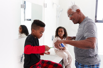 Grandfather In Bathroom Wearing Pajamas Helping Grandchildren To Brush Teeth