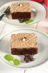 Chocolate bakery style cake brownie  with chocolate cream on top on a white plate and mint leaf and a glass of milk in the background