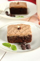 Chocolate bakery style cake brownie  with chocolate cream on top on a white plate and mint leaf and a glass of milk in the background