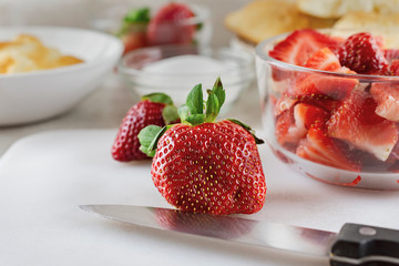 Close up view of a strawberry with knife on a white cutting board.  Bowl of sliced strawberry and full strawberries in the background.  Pastry in the background.