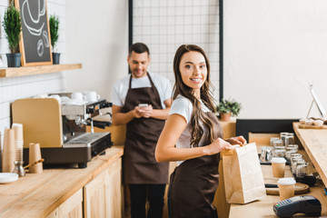 Fototapeta na wymiar cashier with paper bag smiling and barista with smartphone standing behind bar counter in coffee house
