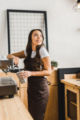 attractive brunette barista in brown apron holding cup and making coffee with machine in coffee house