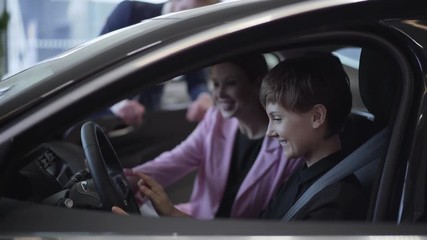 Pretty girl with short hair and her young mother sitting in modern car smiling and examining interior. Salesman standing near the automobile. Woman buying vehicle for her daughter