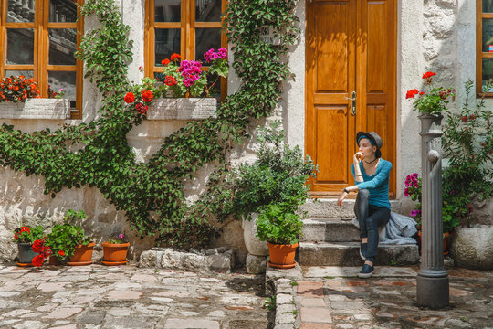 A girl is sitting on the porch of a beautiful old European house shrouded in Vines with a brown wooden door next to her standing water tap.