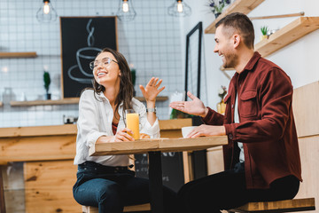 attractive brunette woman with glass of juice and handsome man with cup sitting and laughing at table in coffee house