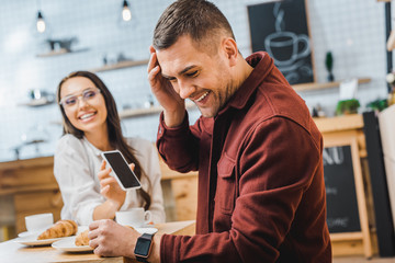 selective focus of handsome man in burgundy shirt sitting at table and laughing wile attractive brunette woman showing smartphone with blank screen in coffee house