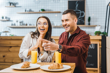 attractive brunette woman and handsome man in burgundy shirt sitting and smiling at table with croissants and glasses of juice in coffee house