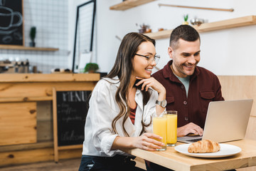 beautiful brunette woman and handsome man sitting at table with juice in glasses and croissants and looking to laptop in coffee house