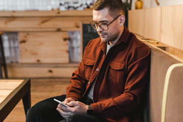 Fototapeta premium handsome man in burgundy shirt, black jeans sitting on couch and looking to smartphone in coffee house