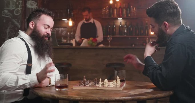 Two stylish men having a conversation over a glass of whiskey and a chess game. In a cozy and stylish pub two bearded man are having a competition.