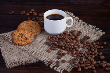 White cup of coffee with coffee grains near and biscuits on linen fabric on the dark vintage background