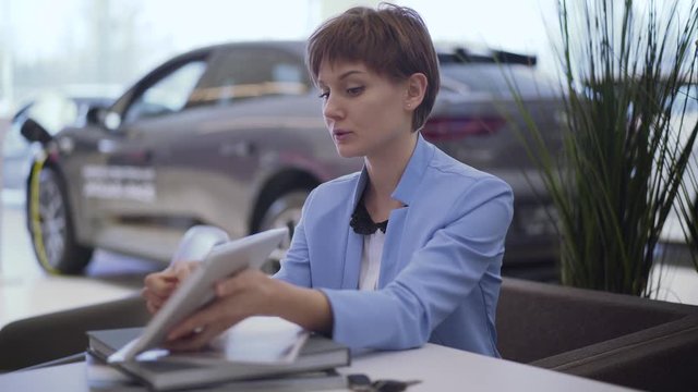 Confident seller woman with short hair in classic blue formal suit shows information to invisible customer on tablet in modern motor show. Blurred car in the background. Saleswoman in car showroom