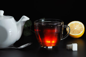 Tea is poured into a transparent cup of white teapot, white sugar and lemon on a black background.