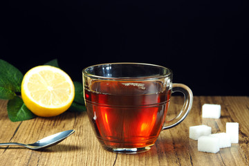 Tea in a transparent cup, lemon and sugar on a wooden table and a black background