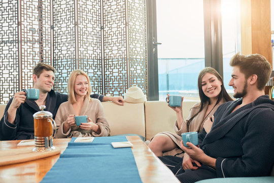 Relaxed diverse friends in bathrobes relaxing at lounge zone of bathhouse, drinking hot tea and sharing positive impressions