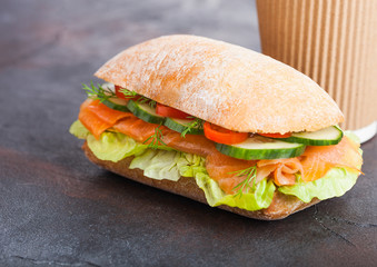 Fresh healthy salmon sandwich with lettuce and cucumber with paper cup of coffee on black stone background. Breakfast snack.