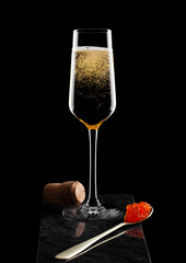 Elegant glass of yellow champagne with red caviar on golden spoon and cork on marble board on black...