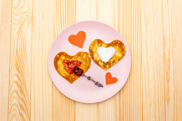 Heart shaped pancakes for romantic breakfast with strawberry jam, silver spoon and paper hearts. Shrovetide (carnival) concept. On wooden background, top view