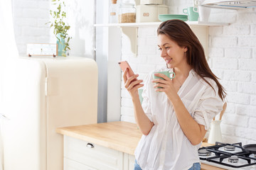 Young woman using smartphone leaning at kitchen table with coffee mug and organizer in a modern home. Smiling woman reading phone message. Brunette happy girl typing a text message