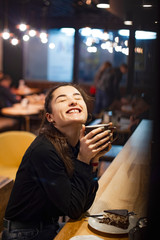 Gorgeous smiling young woman with dark hair eating cake and drinking coffee at a cafeteria in the evening sitting by the window. selective focus, noise effect