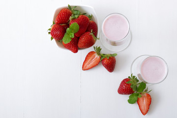 Strawberry smoothie. 2 glasses of strawberry milkshake, porcelain bowl with strawberries embellished with few real strawberries and fresh mint. White wooden background. Free space for text. Top view.