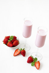 Strawberry smoothie. 2 glasses of strawberry milkshake, porcelain bowl with strawberries embellished with few real strawberries and fresh mint. White wooden background. Free space for text.