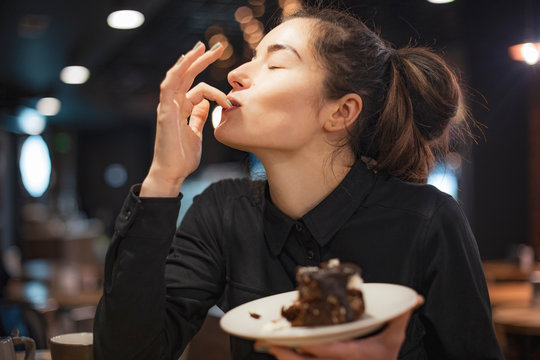Gorgeous smiling young woman with dark hair eating cake and drinking coffee at a cafeteria in the evening sitting by the window. selective focus, noise effect