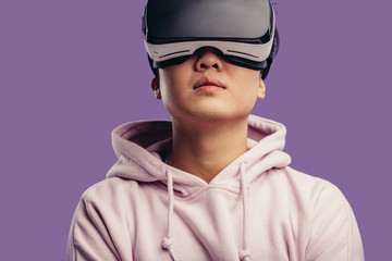 Asian young man dressed in white hoodie using virtual reality headset isolated over violet background