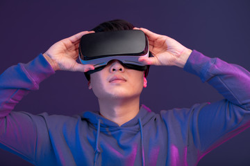 Young Asian man playing VR game in dark blue background. Chinese young hipster wearing VR headset, half body shot. Young generation choose virtual reality relaxation concept.