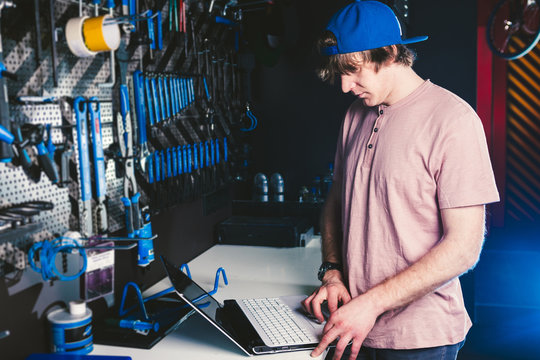 Theme business profession manager and technology. Young stylish Caucasian male owner bicycle shop uses a laptop computer near the stand with the tools for repairing and servicing the bicycle