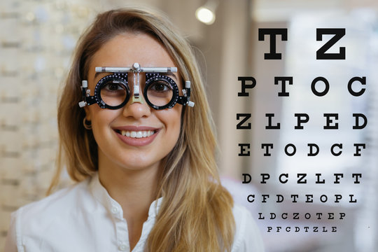 woman wearing trial frame and looking at the camera. beside her visual sight test chart