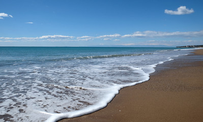 Beautiful landscape with sea, surf with waves with foam runs to long coast with empty sandy beach and high mountains with snow caps on skyline in haze on sunny summer day