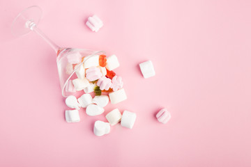 Cocktail glass lying down and from which come out jellybeans, sugar clouds and gummy bears on pink rosewood background